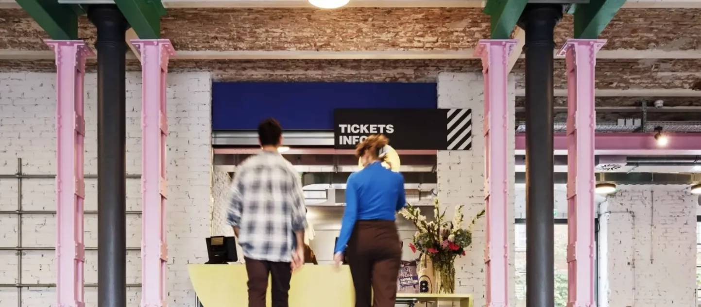 Zwei Personen laufen auf ein Schild mit der Aufschrift 'Tickets, Info' zu in einem Raum mit bunten Stahlträgern und unverputzten Decken