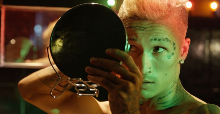 Eine Person mit kurzen Haaren und Tatoos im Gesicht schaut sich in einem Handspiegel an